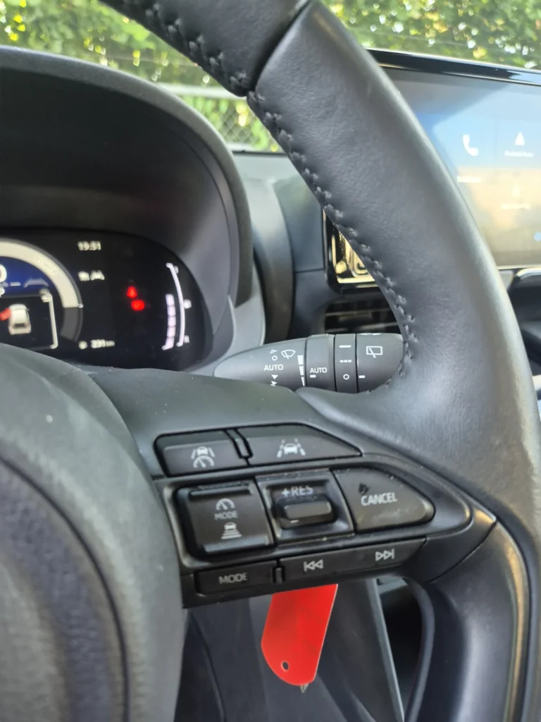 Close-up van een stuurwiel met knoppen en een dashboard op de achtergrond.