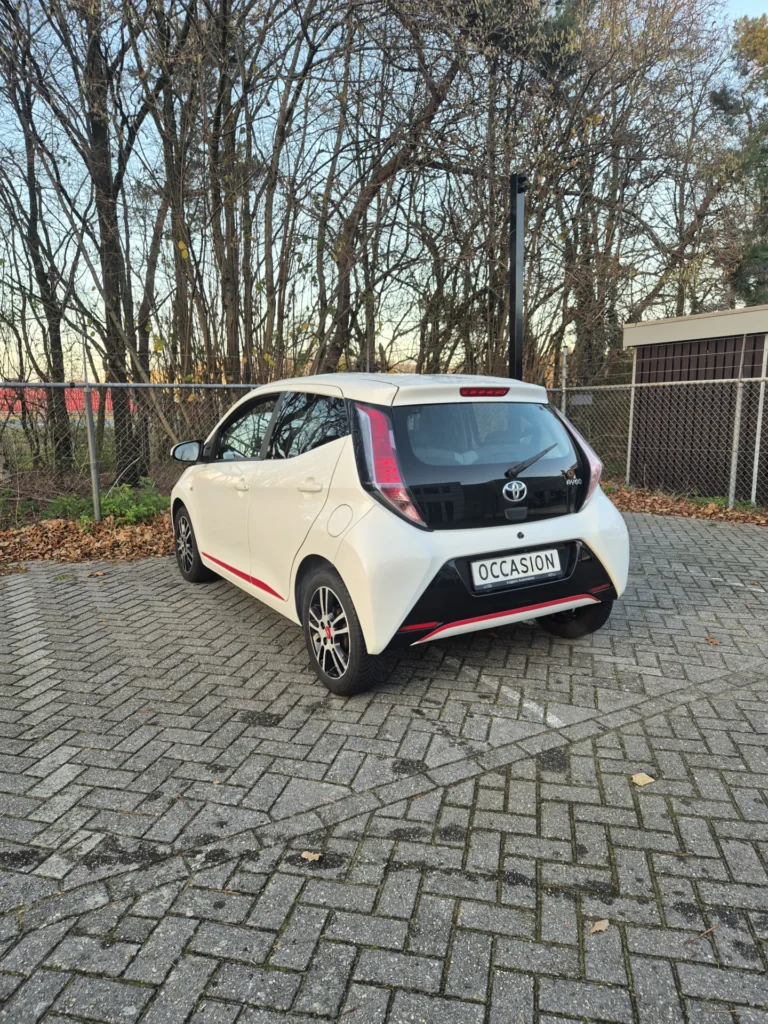 Witte auto met het woord 'Occasion' op de kentekenplaat, geparkeerd op een stenen parkeerplaats.