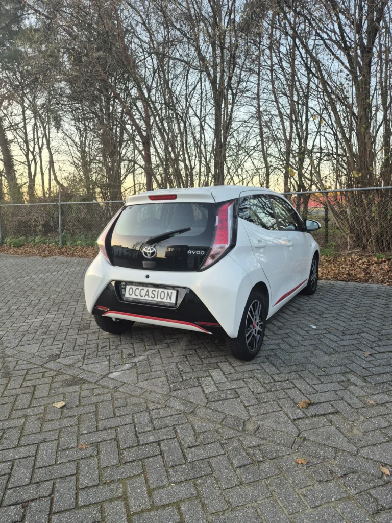 Witte Toyota Aygo geparkeerd op een stenen parkeerplaats naast bomen.